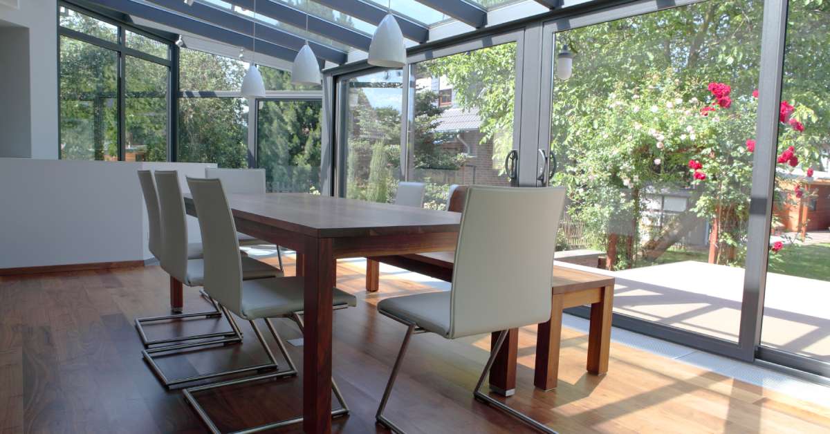 A sunroom in the daytime furnished with a wood dining table, five white dining chairs, and a wood bench facing the backyard.