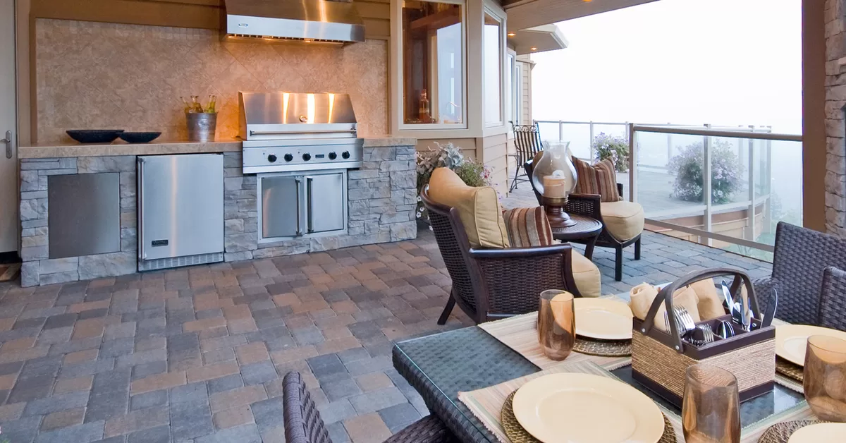 A covered patio with a kitchen area, two chairs, a stone fireplace, and a dining table with four place mats.