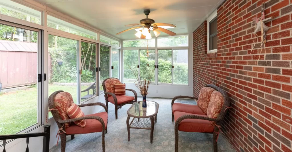 A screened porch with matching wicker furniture and a glass coffee table adjoined to a red brick house.