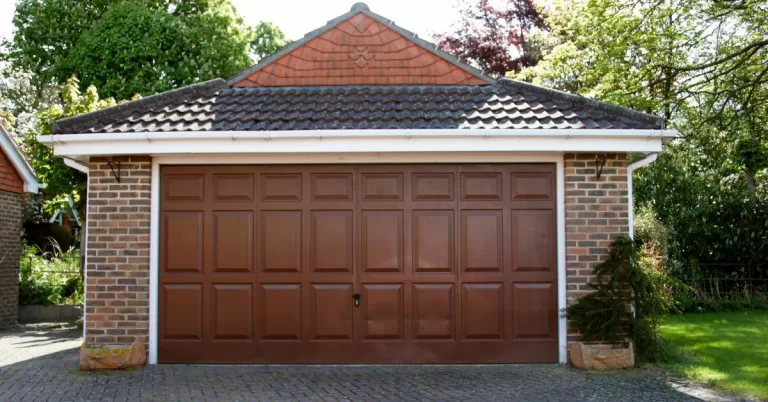 A detached garage with a brick exterior, shingle roof, and chocolate brown door with several trees in the background.