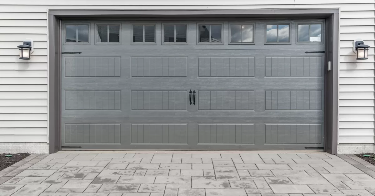 A grey garage door on a white house with a stone driveway. The door features a row of windows and black handles.