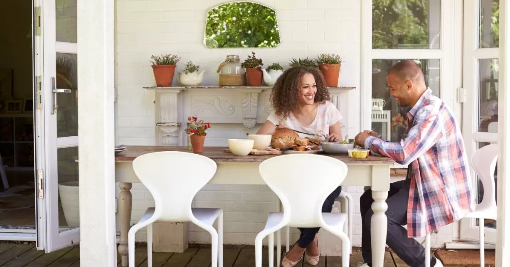 A couple sit at a dining table eating a meal with a loaf of bread and salad bowl outside on their back porch.