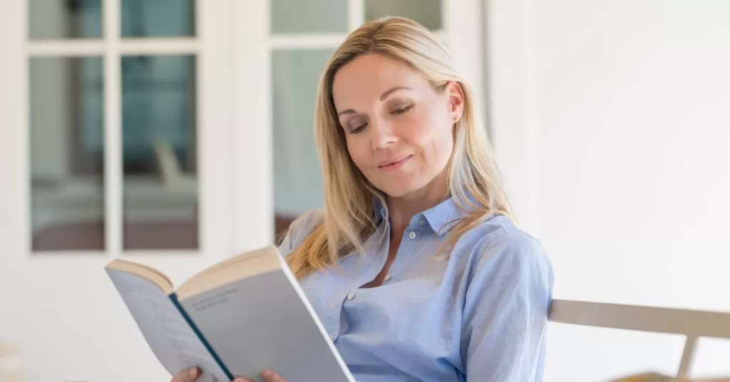 A middle-aged woman wearing a blue buttoned-down shirt is reading a white paperback book on a porch.