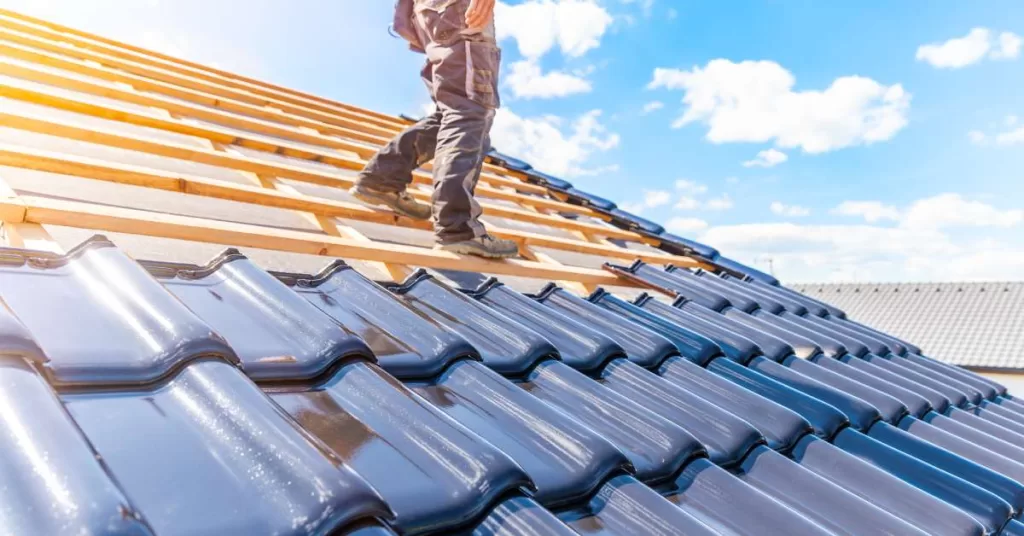 A roofer stands on the exposed beams of a residential roof under construction with some dark ceramic tile laid.