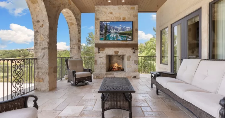 A luxury porch has stone arch walls, a stone fireplace with a mounted TV, and matching outdoor furniture.