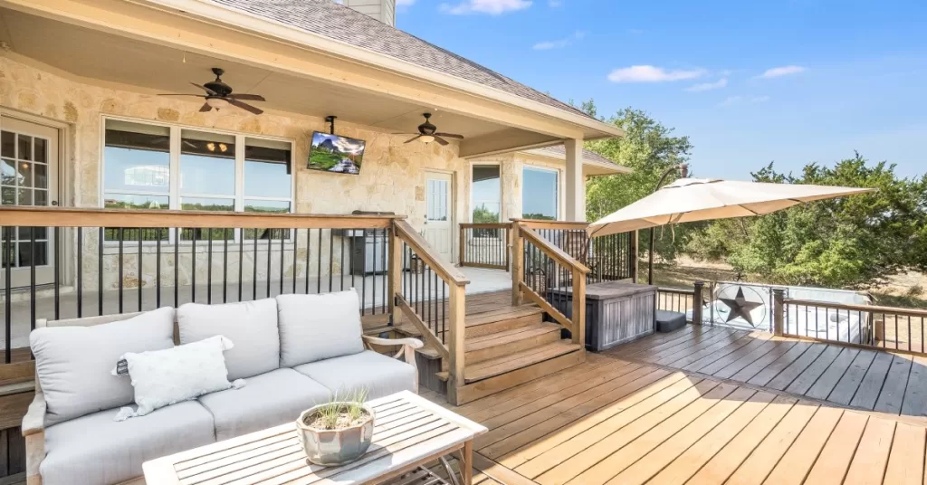 A backyard porch has steps leading to a large, uncovered wooden deck with outdoor furniture and a big umbrella.