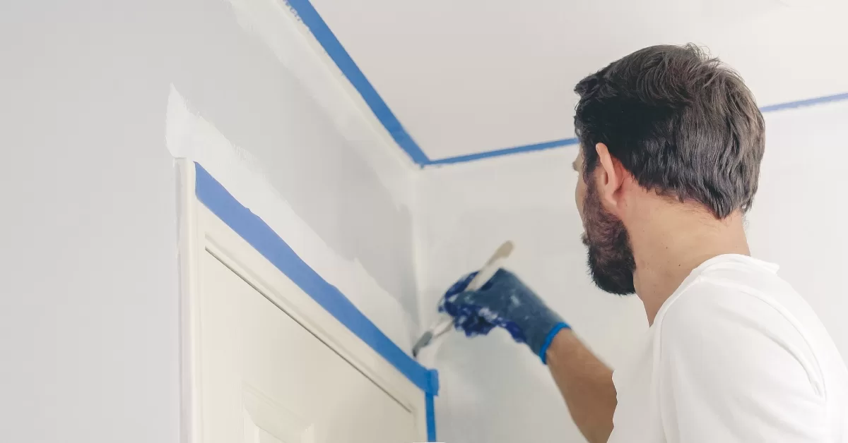 A man wearing a white t-shirt. He is painting a wall white above a white door frame separated by blue painter's tape.