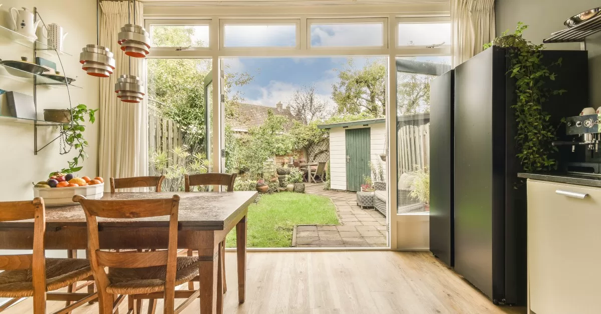 A kitchen with a wood dining table and large glass doors open outward to a backyard with a white and green shed.