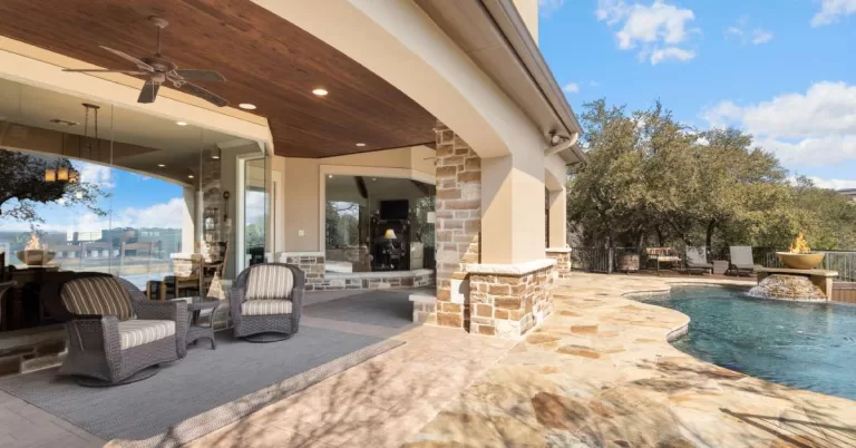 A backyard covered patio made from natural stone next to a pool. Two chairs and a rug sit in the shade.