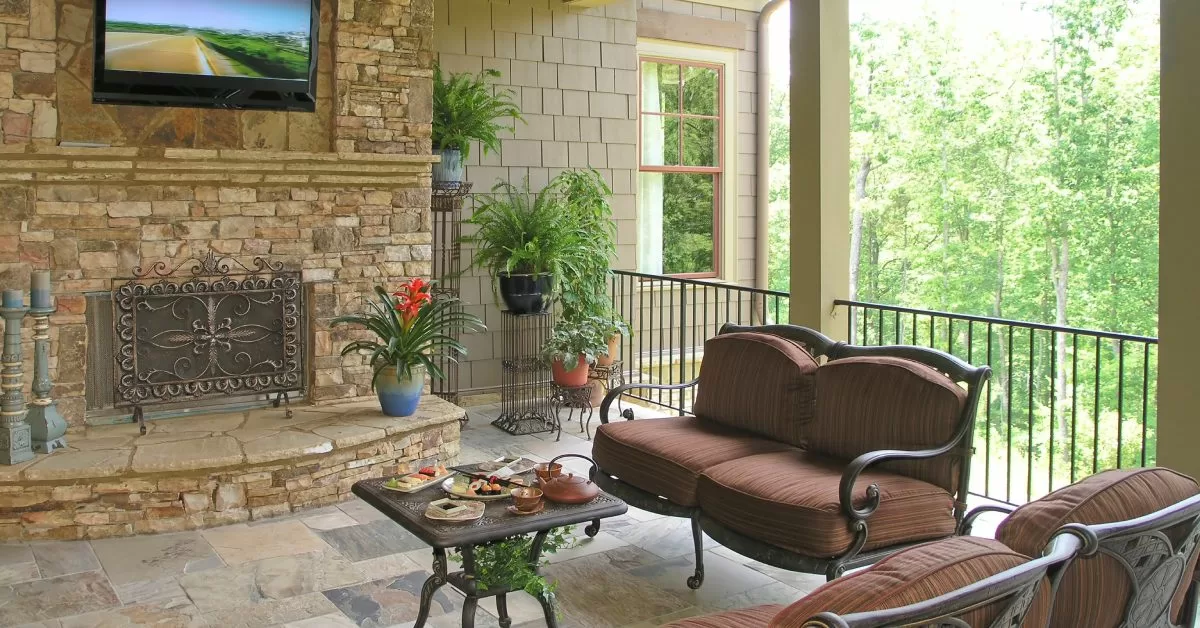 A covered patio with a built-in TV over a stone fireplace. Two matching brown couches face the TV and a square table.