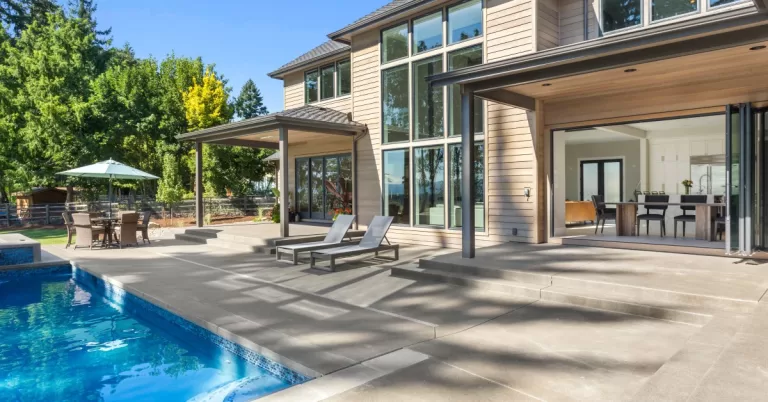 The backyard of a luxury two-story home with tall windows. The patio features lounge chairs, a dining set, and a pool.