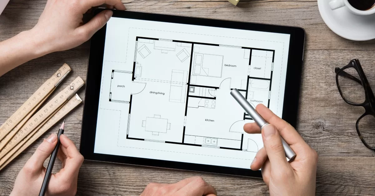 Two sets of hands gesture to a tablet displaying a home floorplan on a wood surface near a folding ruler and a cup of coffee.