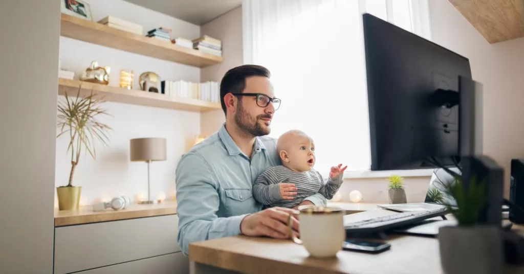 A man wearing a denim shirt and glasses sits at his home office desk while holding a baby on his lap.