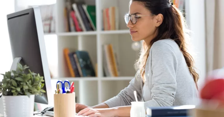 A woman wearing a cardigan and glasses sits at a desk while typing on a keyboard in her home office.