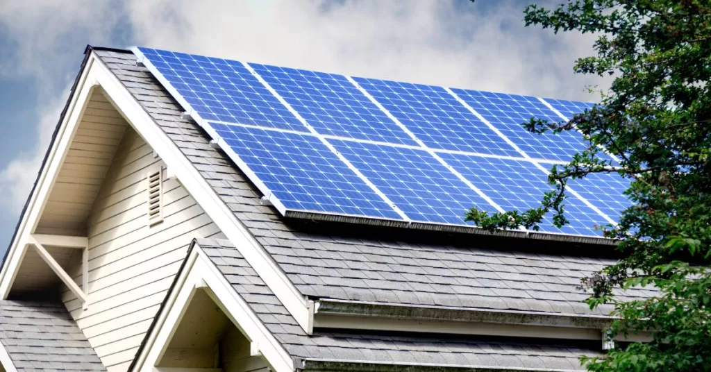 A residential home that features an asphalt shingle roof and several solar panels reflecting the blue, cloudy sky.