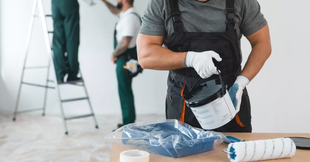 A man wearing coveralls pours a can of paint into a paint tray on a table. Two painters talk in the background.