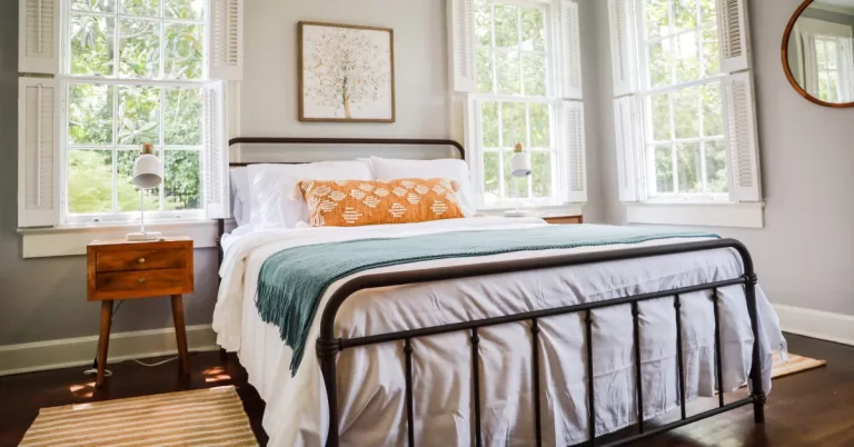 Sunlight streams through three windows into a guest bedroom featuring a queen mattress and two matching side tables.