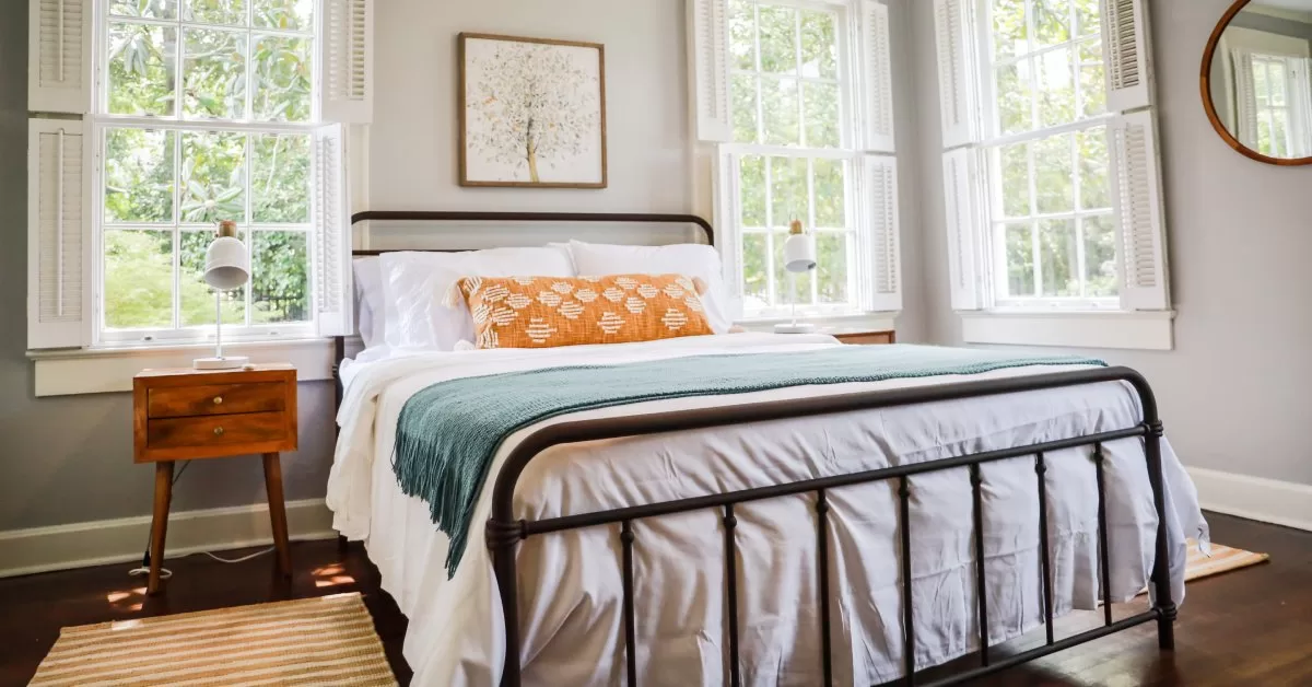 Sunlight streams through three windows into a guest bedroom featuring a queen mattress and two matching side tables.