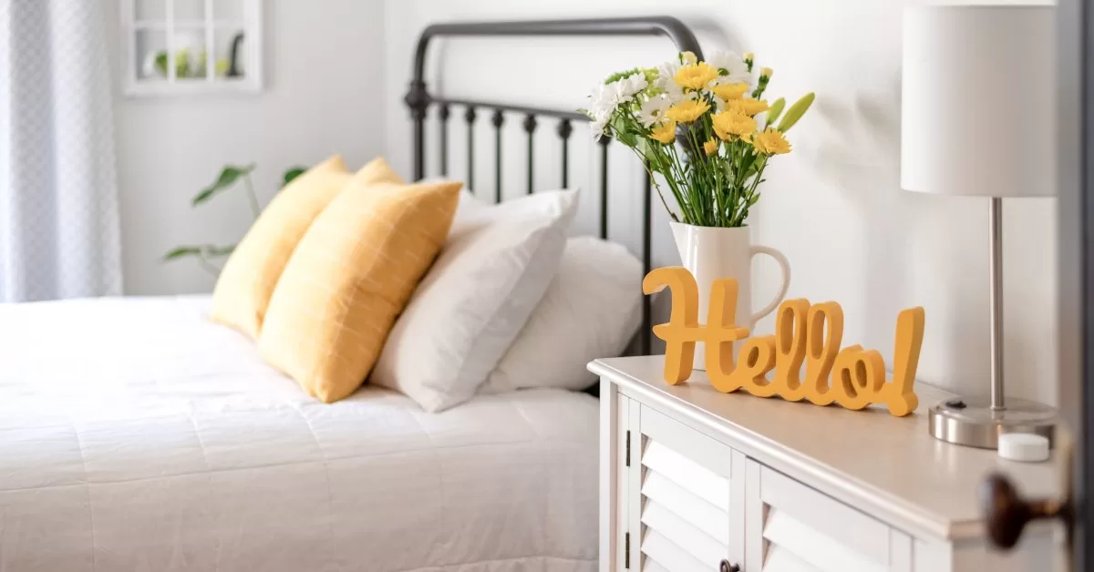 A guest bedroom with white and yellow bedding. A sign reading "hello!" in yellow cursive font sits on a white side table.