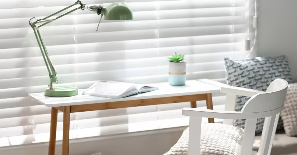 A small white desk in a bedroom with a white chair with a green lamp facing a window with light shining through the blinds.