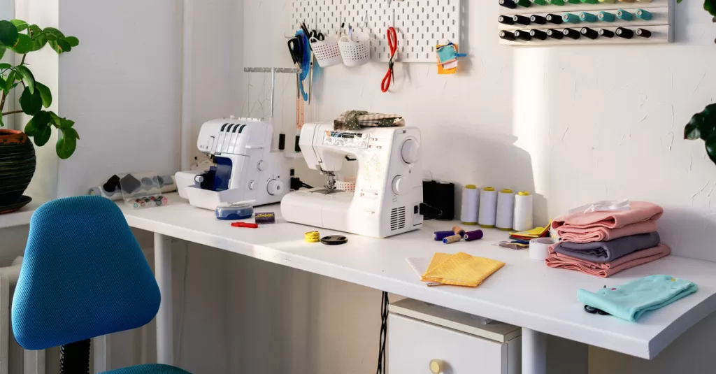 A sewing workspace with a serger and sewing machine. A pegboard and thread spool display hang on the wall.