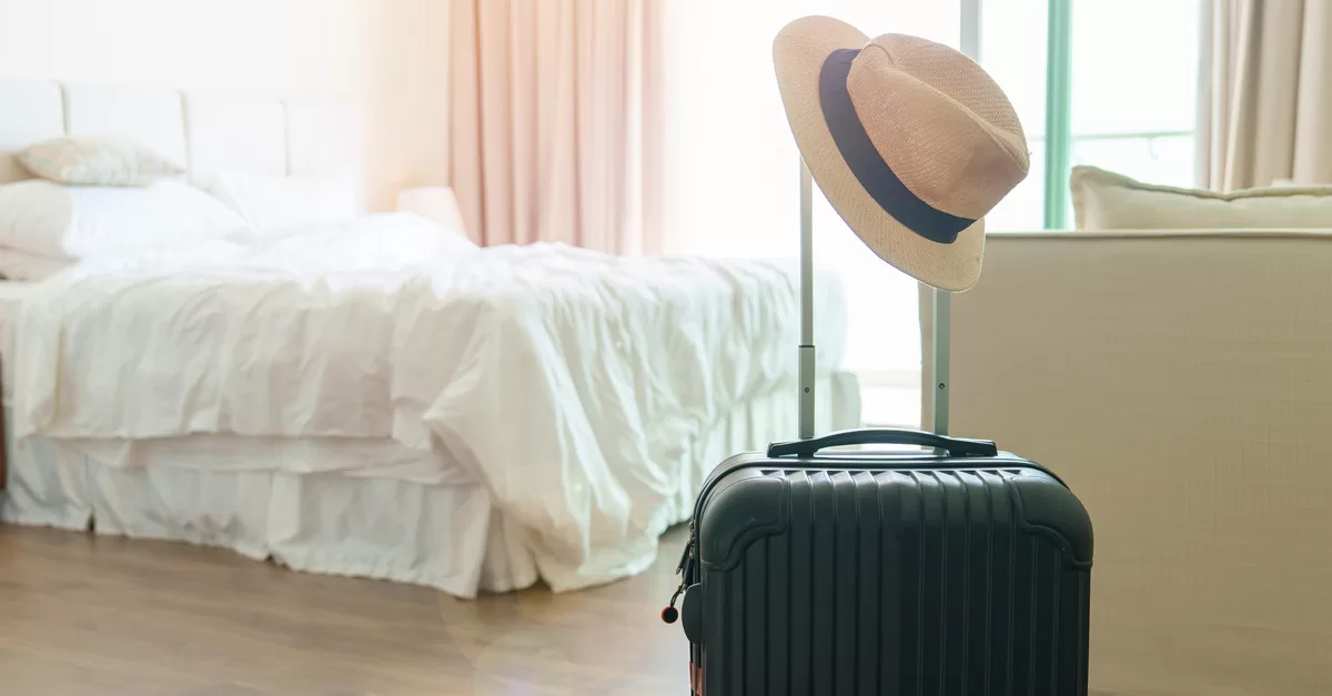 A black suitcase with a woven hat sitting on the handle in a guest bedroom with a white bed and cream couch.