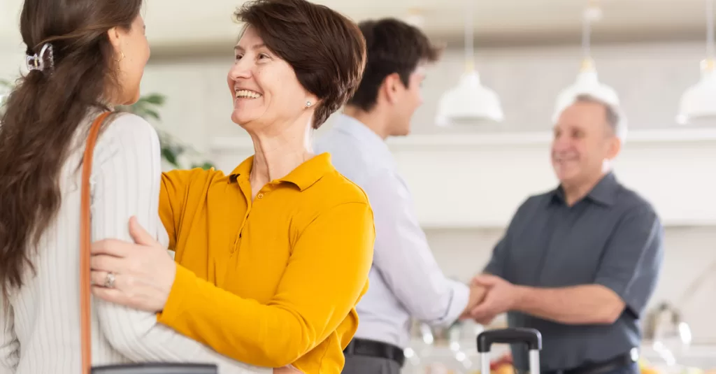 Parents welcome their adult daughter and son into their home. Both carry suitcases while greeting their parents.