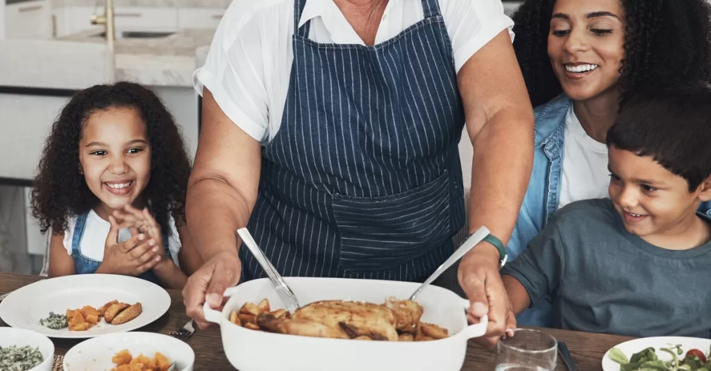 A grandmother in an apron places a dish of cooked chicken on the dining room table, where a woman and children are seated.