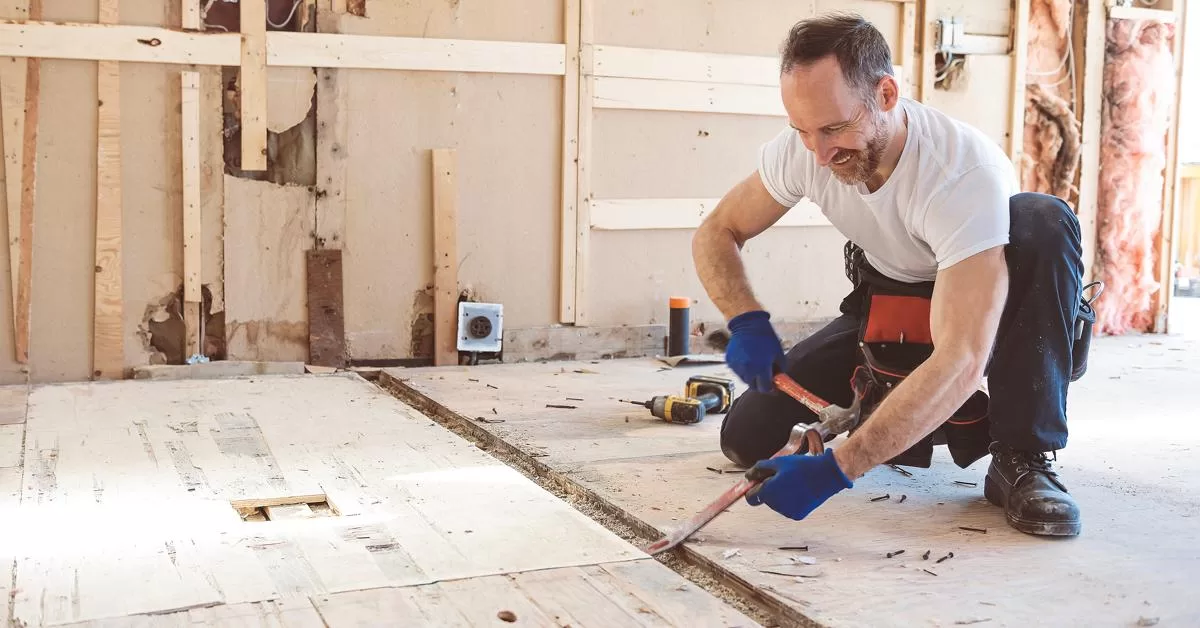 A male contractor uses a crowbar and hammer to pry a base layer of flooring in a home with exposed framing.