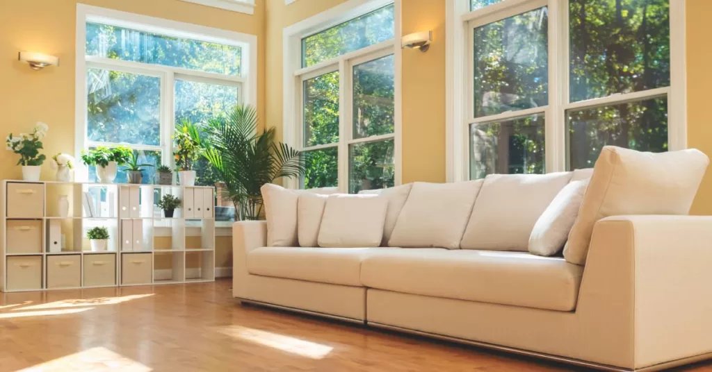 A home common area with a cream couch against a wall with several large windows facing a forest with sunlight shining through.